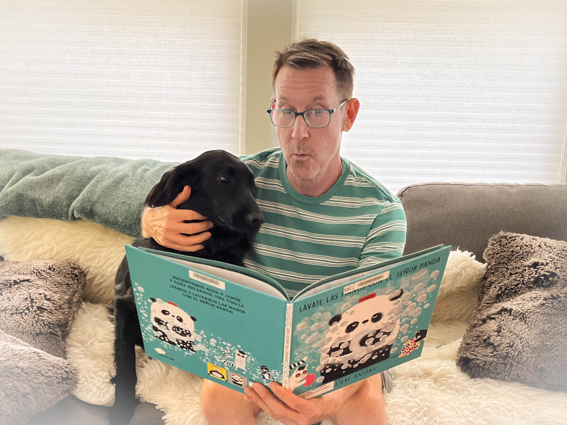 A man reading a book to a black dog on a couch.