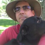 Man wearing a hat and sunglasses holding a black dog outdoors.