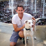 Man kneeling with a dog by a fountain.
